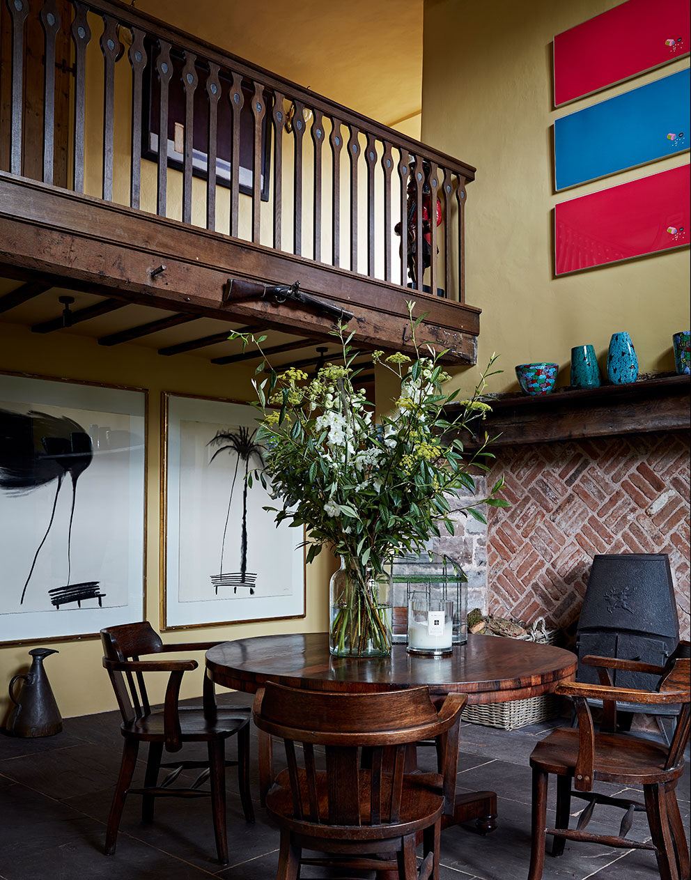 A round wooden table in a double-height dining room with herringbone brick fireplace and contrasting modern art.