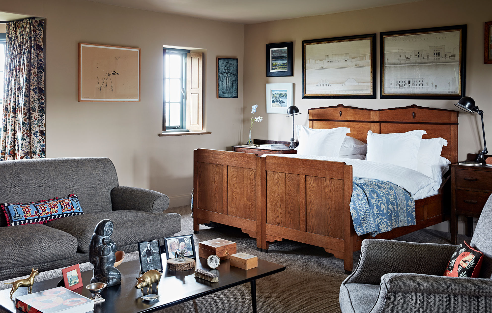 Bedroom of a rural home designed by Godrich with a vintage wooden bed, soft natural walls covered in art and a comfortable seating area.