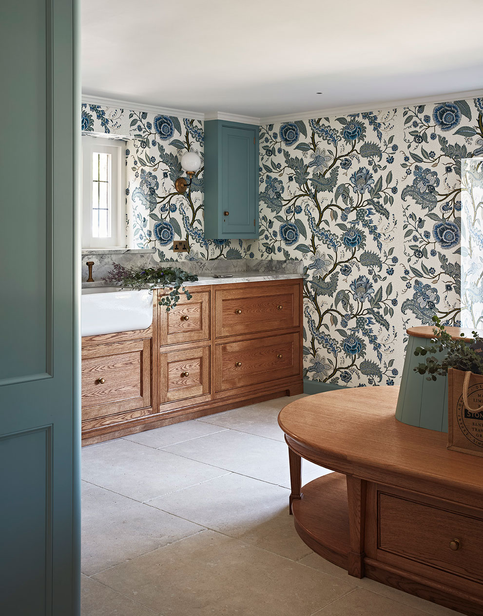 Boot room of a manor house with a matching teal colourway in the wallpaper and cupboards, contrasting beautifully with the wooden drawers and white marble countertop.