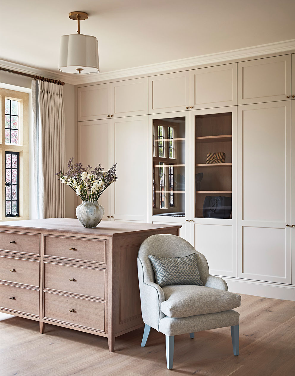 Fitted cupboards in a muted tone in a room with wooden floors, a free-standing island cupboard and a grey upholstered armchair.