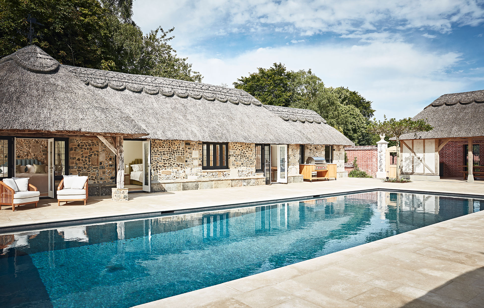 Stunning outdoor swimming pool surrounded by low stone uildings with thatched roofs, all with interior design by Godrich Interiors.