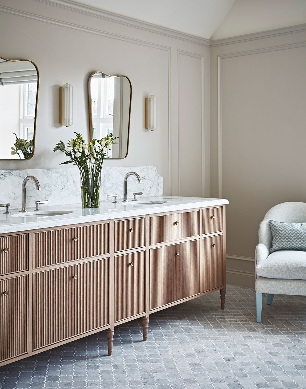 Light-filled room with a muted palette with two sinks, mirrors and a grey upholstered armchair.
